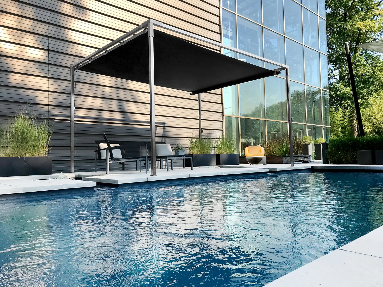 SHADE CUBE als Poolbeschattung vor einer Glasfassade: freistehender Edelstahl-Rahmen mit dunkler Segelmembrane