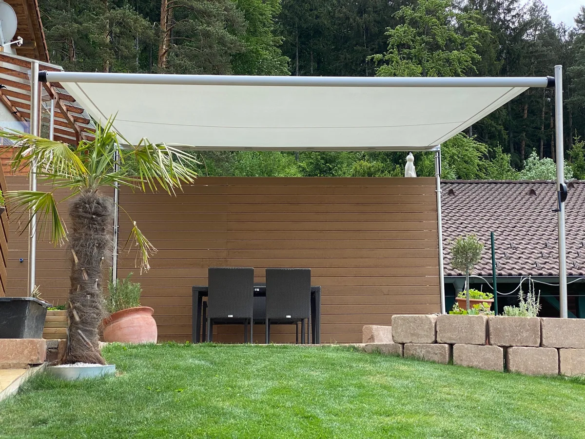 SHADE FRAME über einem Sitzplatz im Garten vor einer Holzwand, Segelmembrane weit ausgefahren