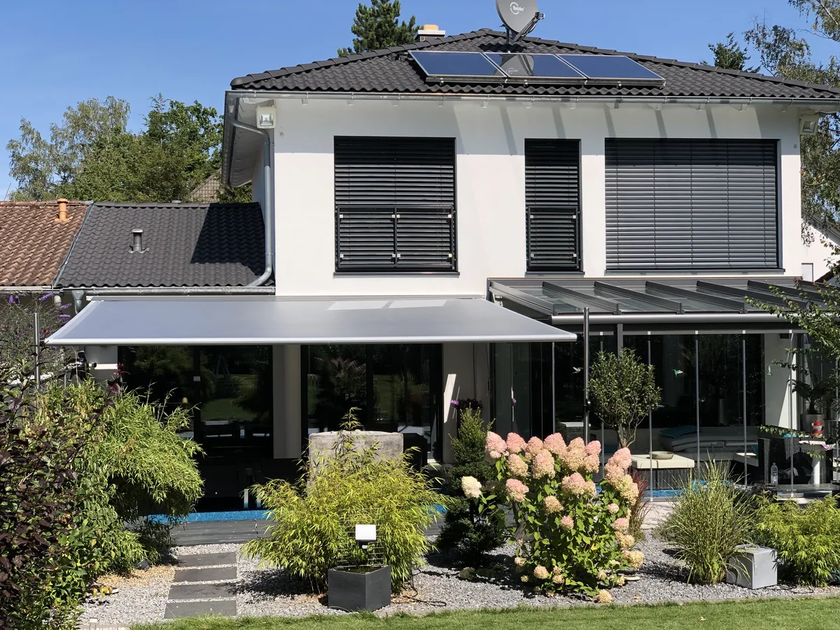 SHADE INOX an der Hausfassade: ausgefahrenes Sonnensegel über der Terrasse neben dem Glas-Wintergarten