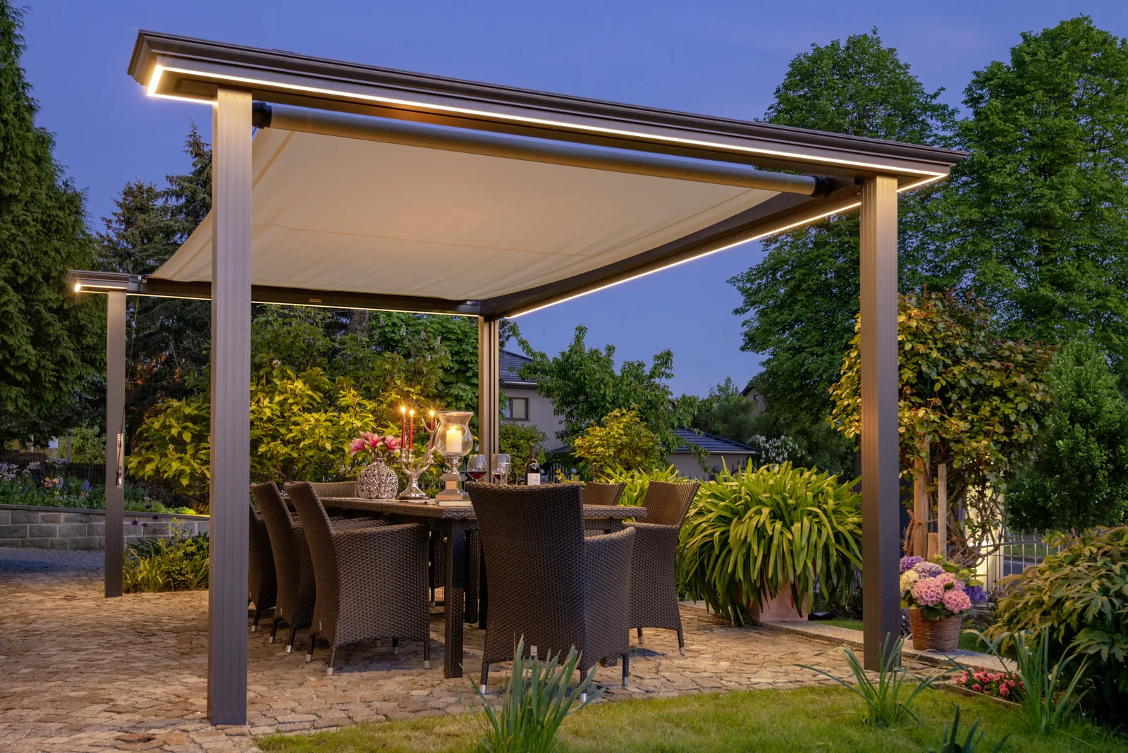 SHADE STRUCTURE am Abend: Sonnensegel unter einer Pergola über dem gedeckten Terrassentisch, Rahmen mit LED-Beleuchtung